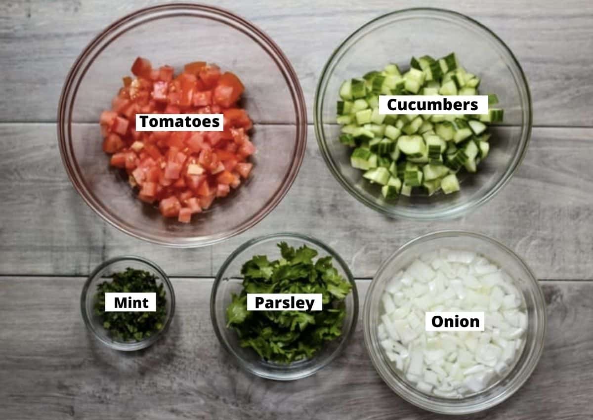 Chopped tomato, cucumber, mint, parsley, and onion in glass bowls. 