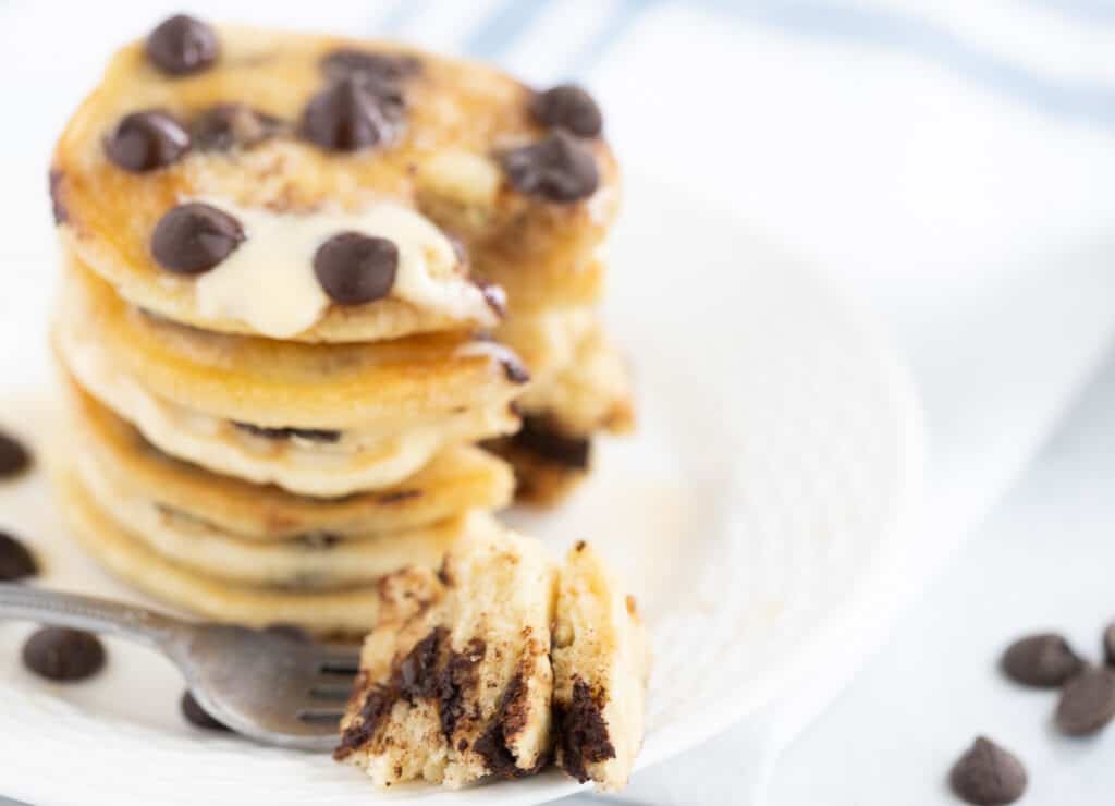 Chocolate chip pancake bite on fork beside stack of pancakes on plate.