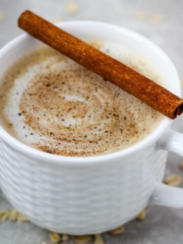 Oat milk latte in small white cup topped with cinnamon stick.