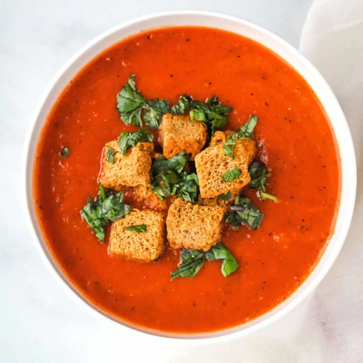 Bowl of creamy vegan tomato soup with croutons and fresh herbs.
