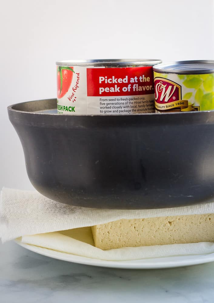 Tofu pressing between paper towels, with large saucepan filled with cans on top. 