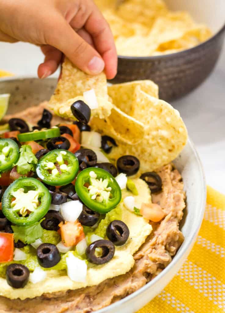 vegan 7 layer dip in white bowl topped with black olives vegan cheese and vegetables