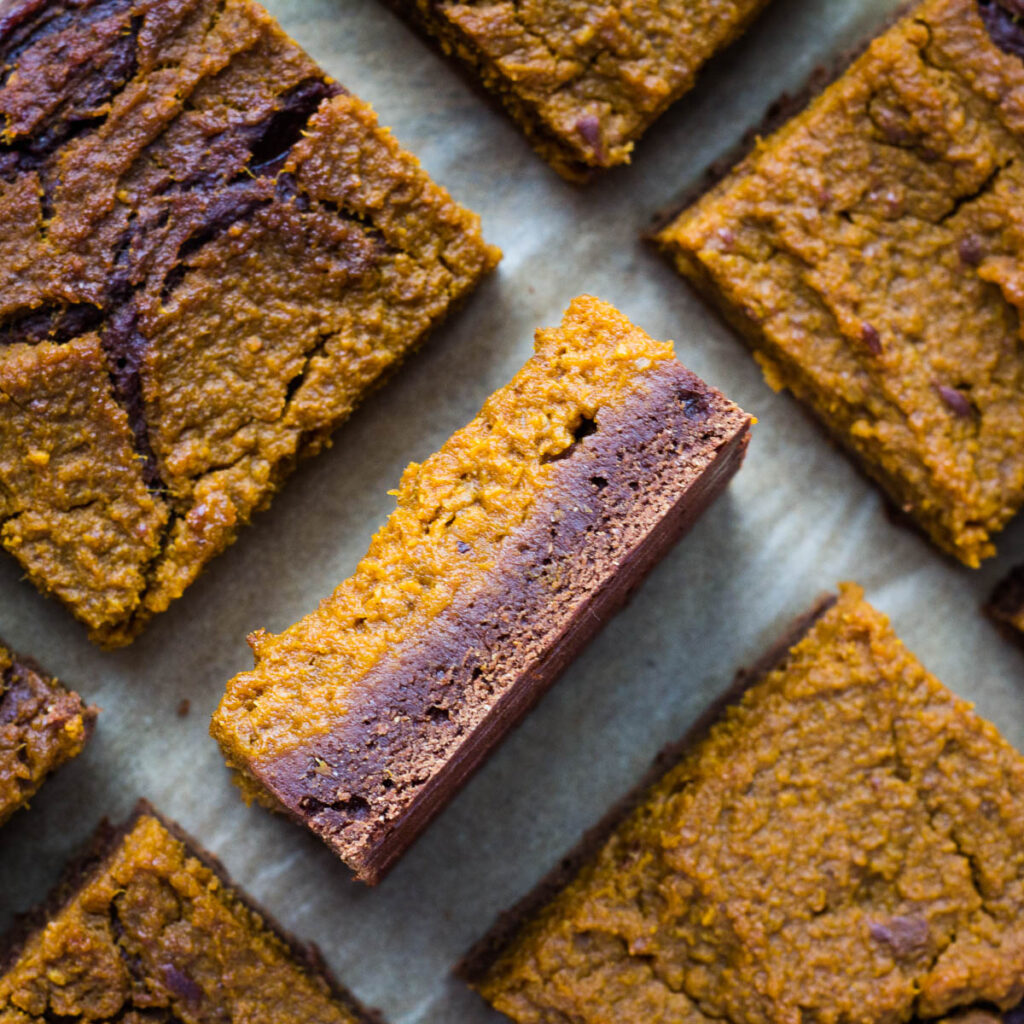 Vegan pumpkin brownies on parchment paper with one brownie on its side to show the pumpkin and brownie layers.