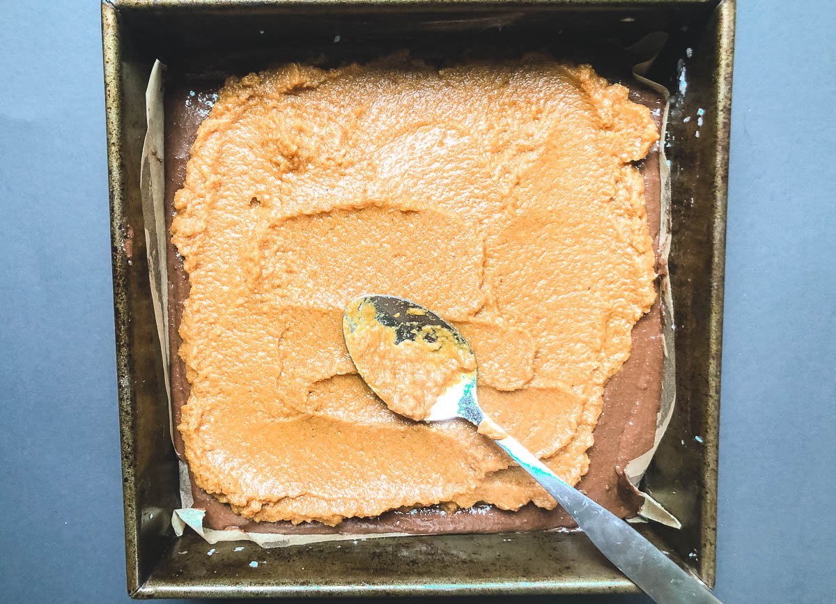 Spreading pumpkin puree over brownie batter with spoon. 