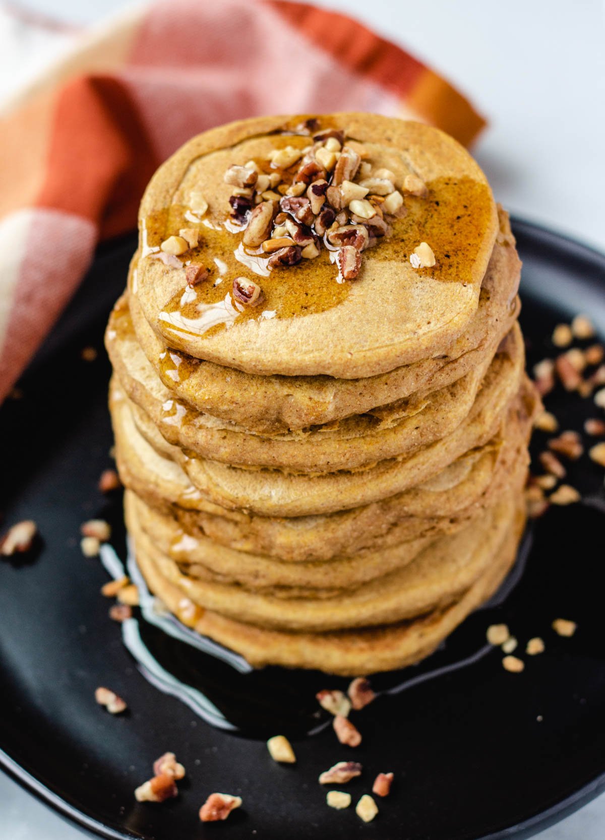 Stack of vegan pumpkin pancakes topped with chopped nuts and maple syrup on black plate.