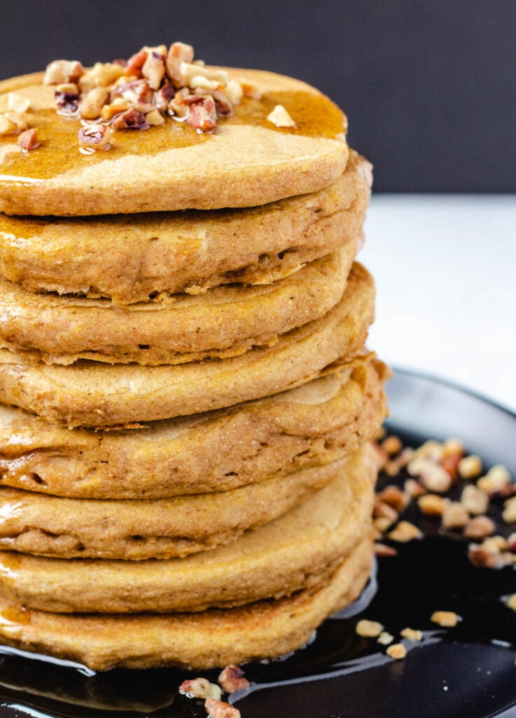 Close up of stack of pancakes topped with nuts and syrup on black plate.