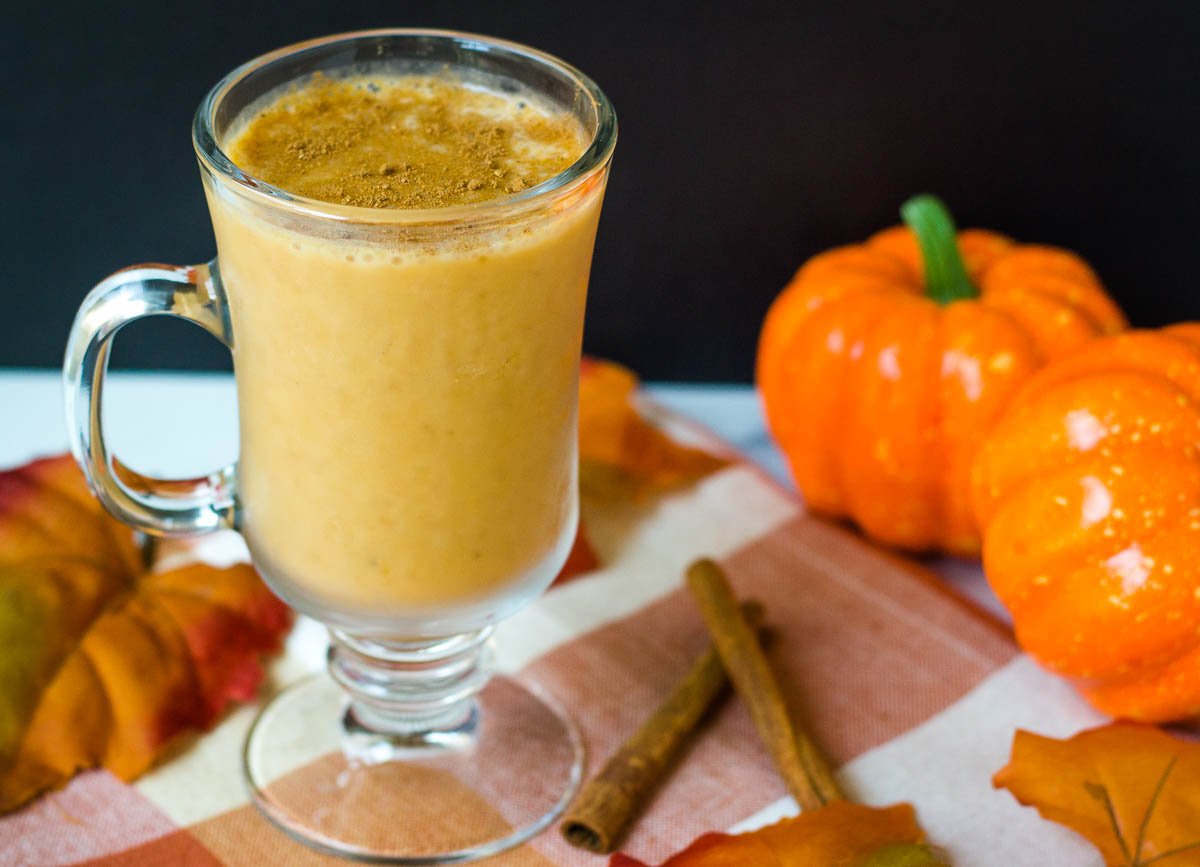 Pumpkin smoothie in glass surrounded by decorative pumpkins and fall leaves.