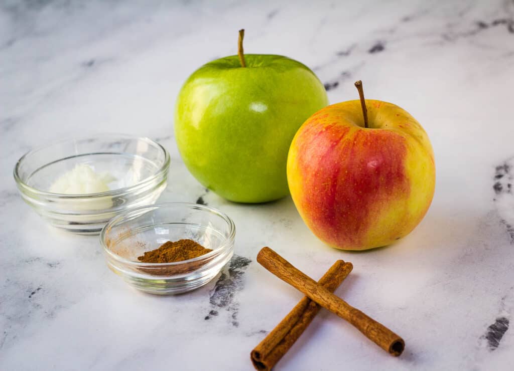 two apples, cinnamon, cinnamon sticks, and coconut oil on counter
