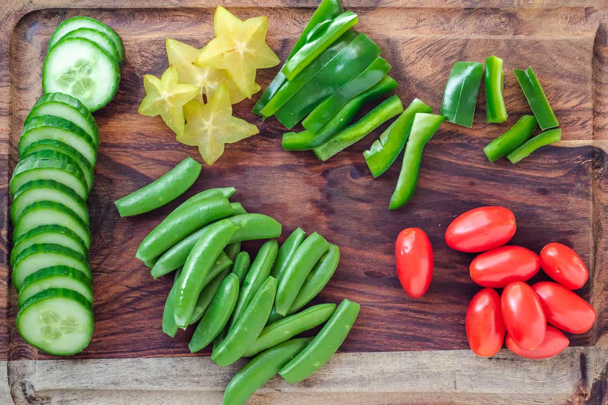 prepared vegetables on cutting board: sliced cucumber, chopped green peppers, chopped starfruit, snap peas, and grape tomatoes