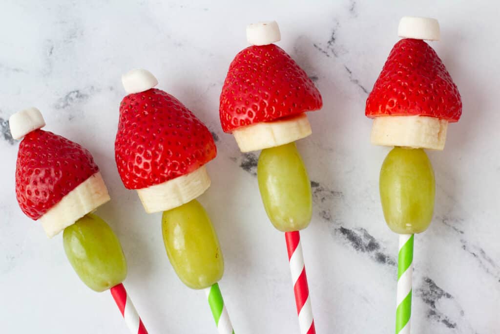 four grinch fruit kabobs made of green grapes, banana, strawberry and marshmallow