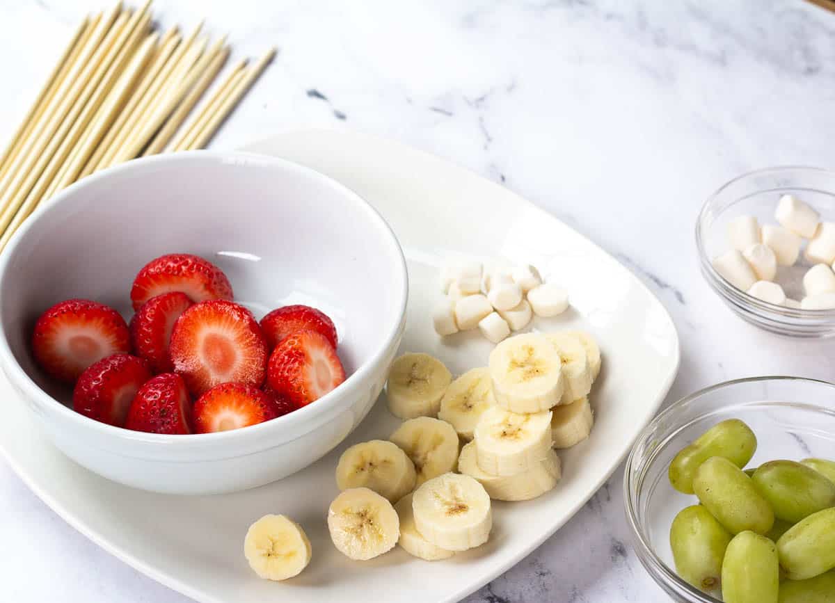 Sliced strawberries, sliced banana, green grapes, on plate, with marshmallows in the background.
