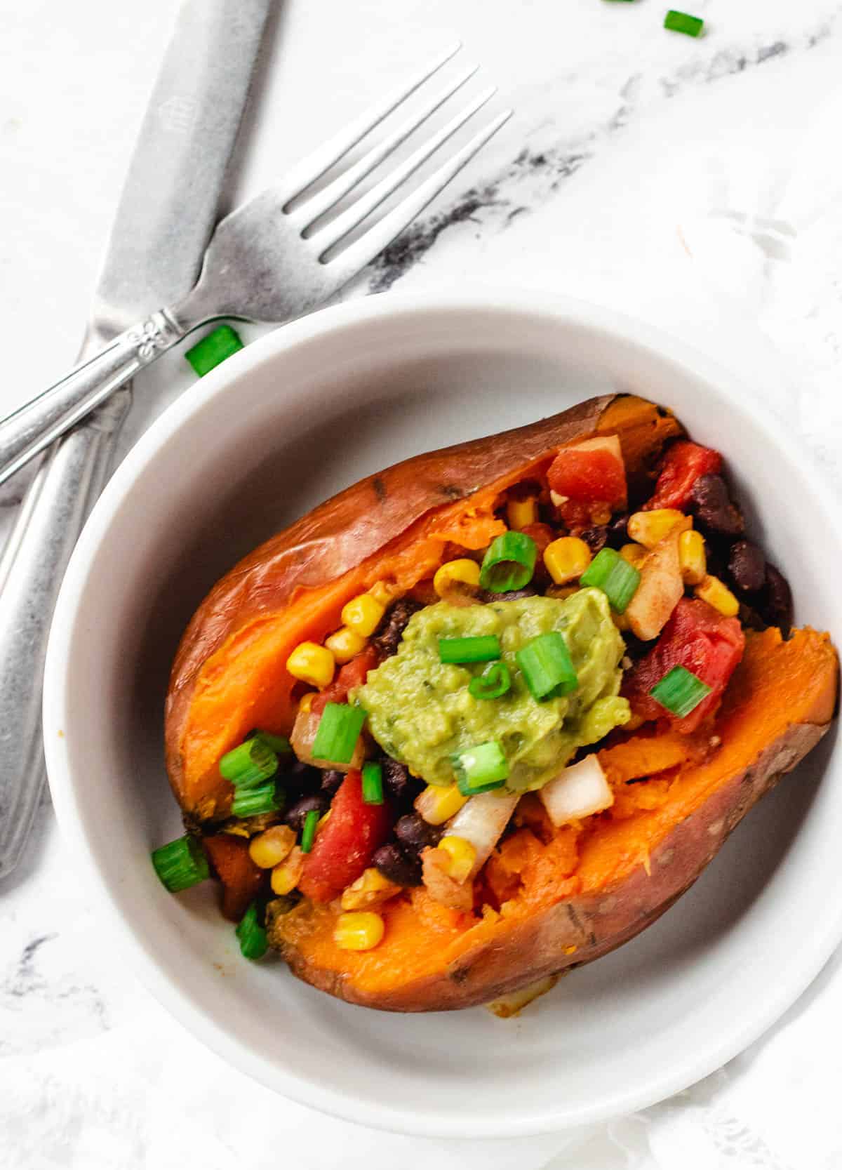 vegan stuffed sweet potato in white bowl topped with corn, beans, tomatoes and guacamole