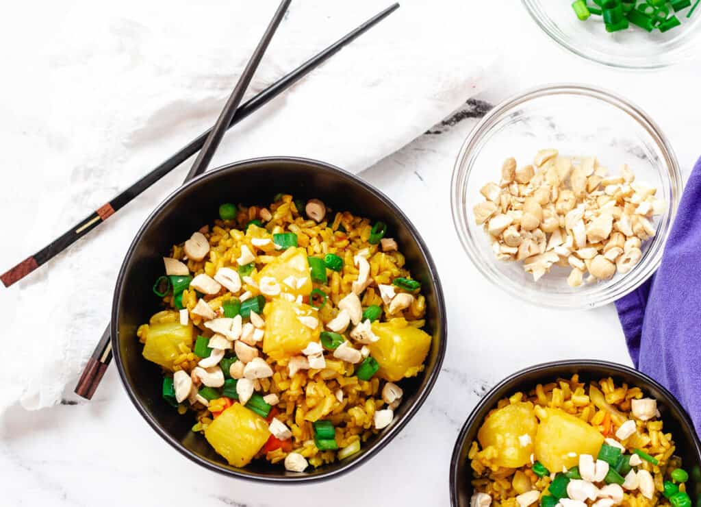 pineapple fried rice in black bowls with chopsticks and a side of chopped cashews
