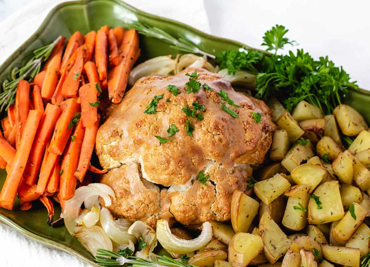 vegan roast dinner on platter. cauliflower, potatoes, carrots, onions, and fresh herbs