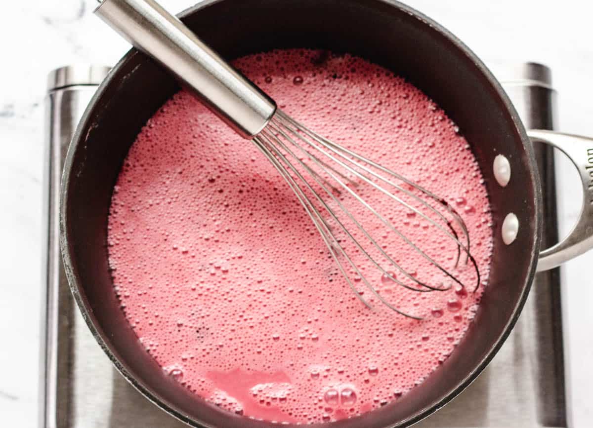 Aaucepan with pink foamy milk and whisk.