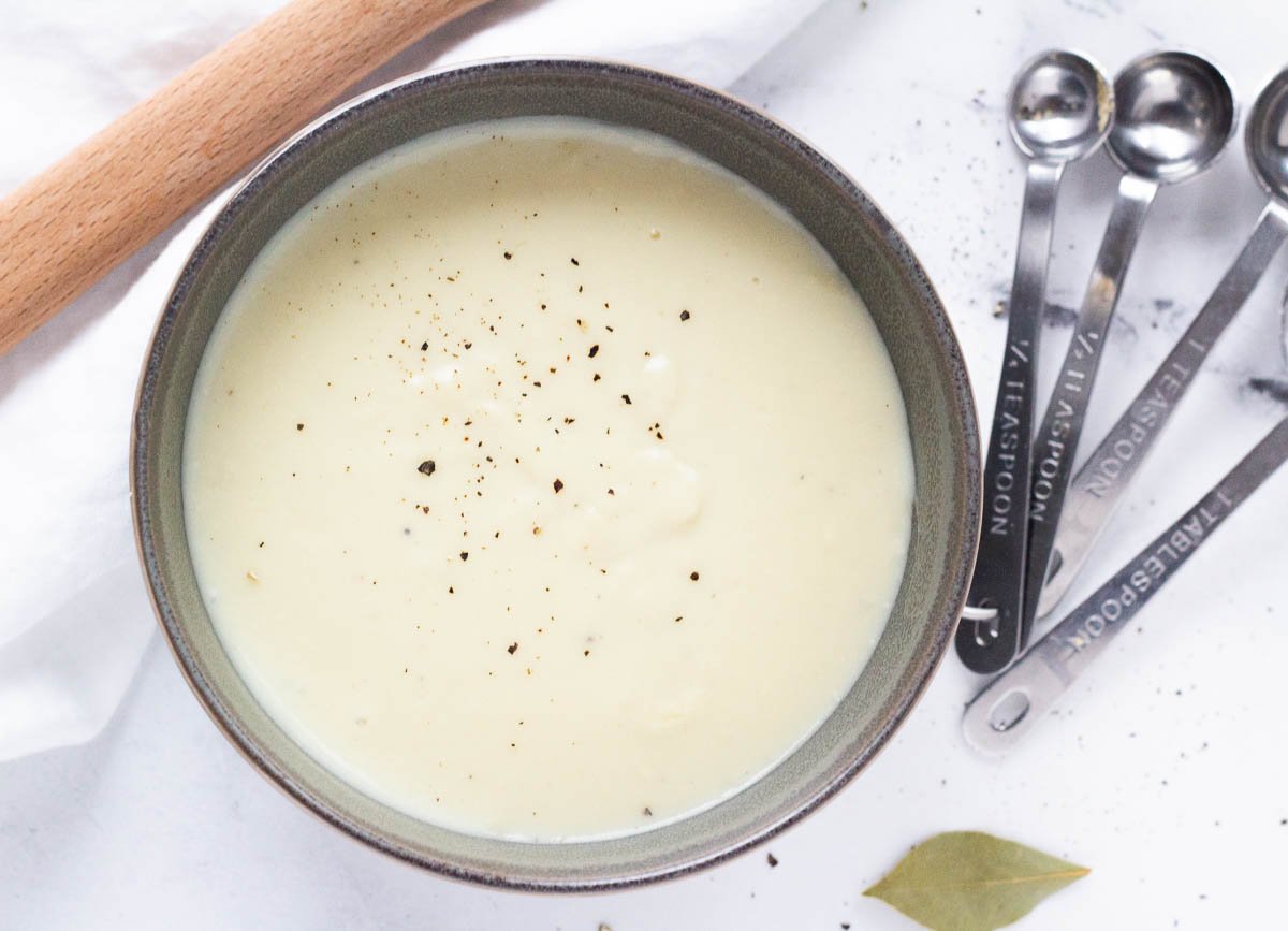 Creamy vegan white sauce topped with black pepper beside measuring spoons and bay leaf.
