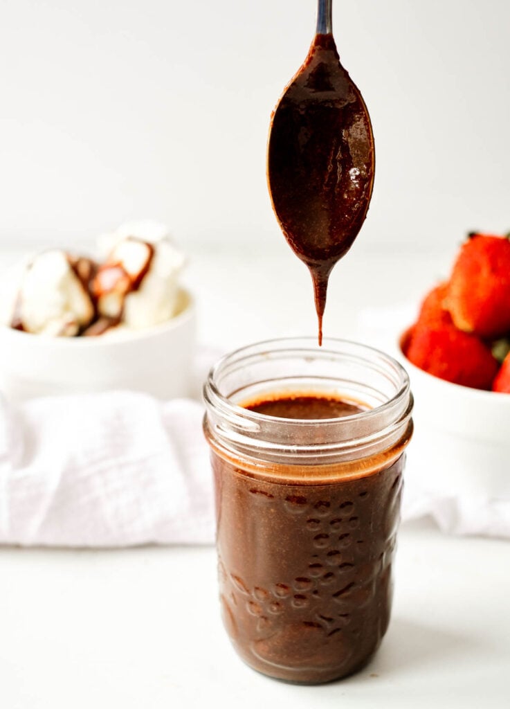 vegan chocolate sauce in mason jar with spoon dipped in sauce hanging over the jar with a drip coming down. Ice cream and strawberries in the background.