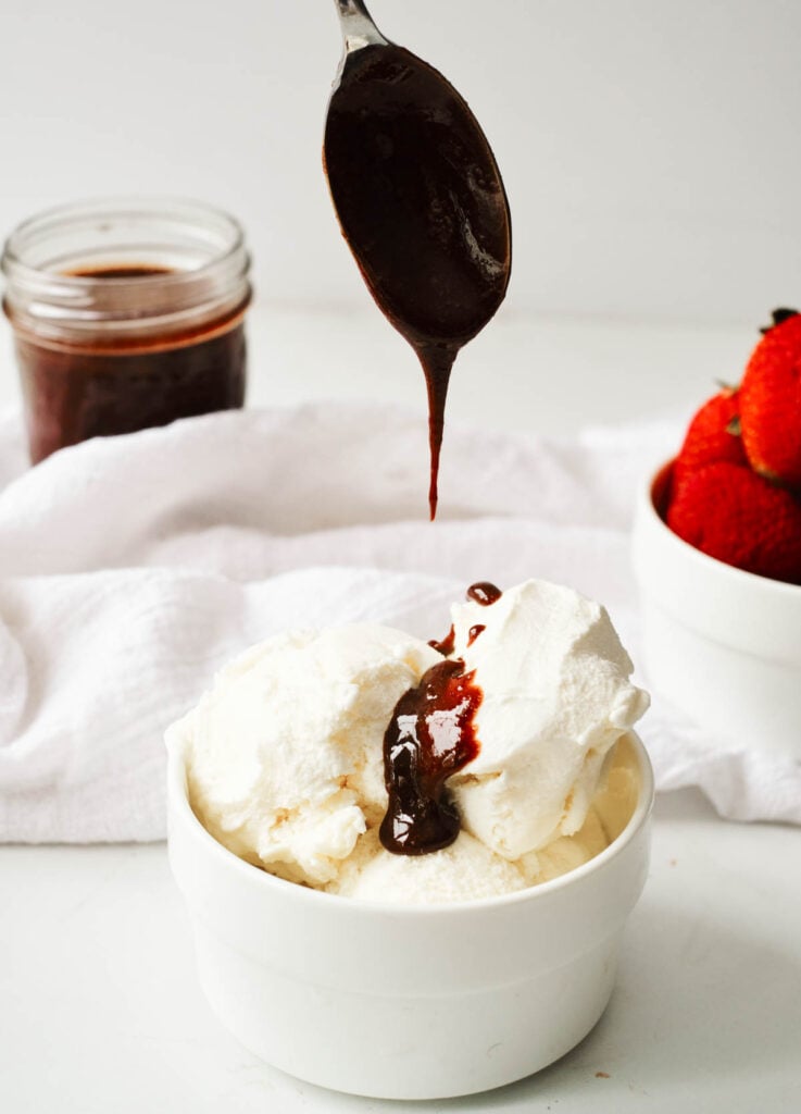 spoon drizzling chocolate sauce over vanilla ice cream in white bowl, with mason jar filled with chocolate sauce, and bowl full of strawberries in the background