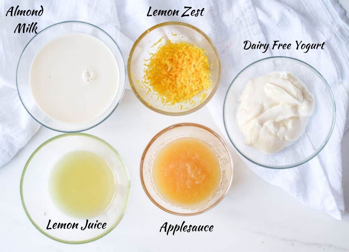 Vegan lemon loaf wet ingredients: almond milk, lemon zest, lemon juice, applesauce, and dairy-free yogurt in glass bowls.