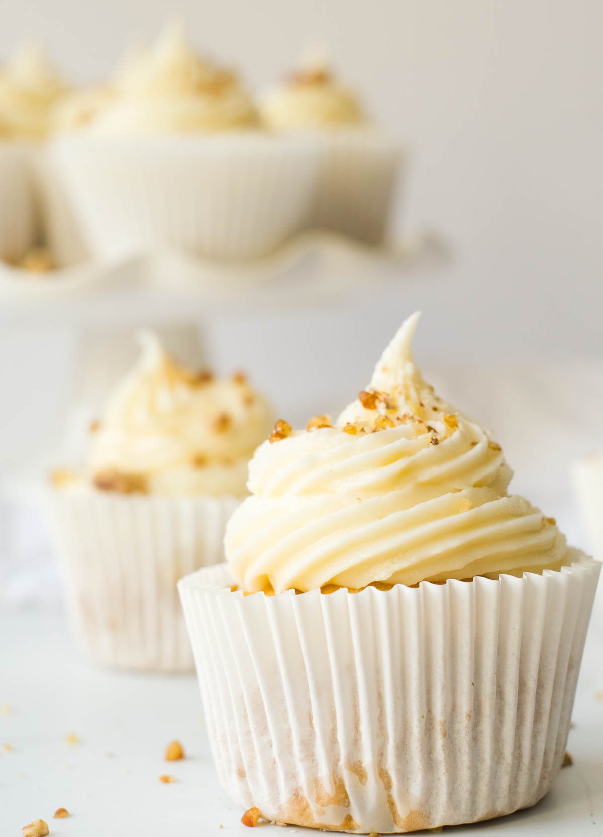 Close up of carrot cake cupcakes on white counter topped with cream cheese and chopped walnuts.