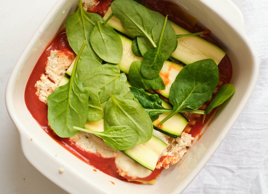 layers of lasagna with layers of marinara sauce, gluten-free lasagna noodles, cashew ricotta, vegan bechamel, sliced zucchini, and spinach