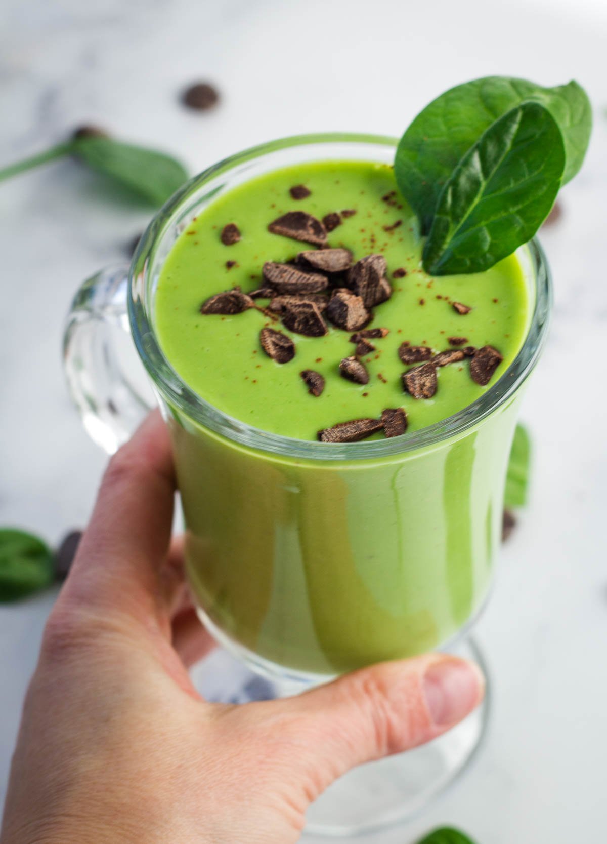 hand picking up glass of green shake topped with chocolate chips and spinach leaves