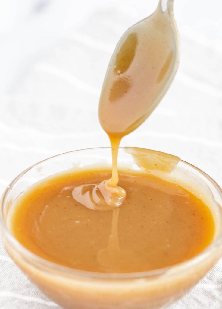spoon dipped in vegan caramel sauce drizzling into small bowl filled with caramel sauce