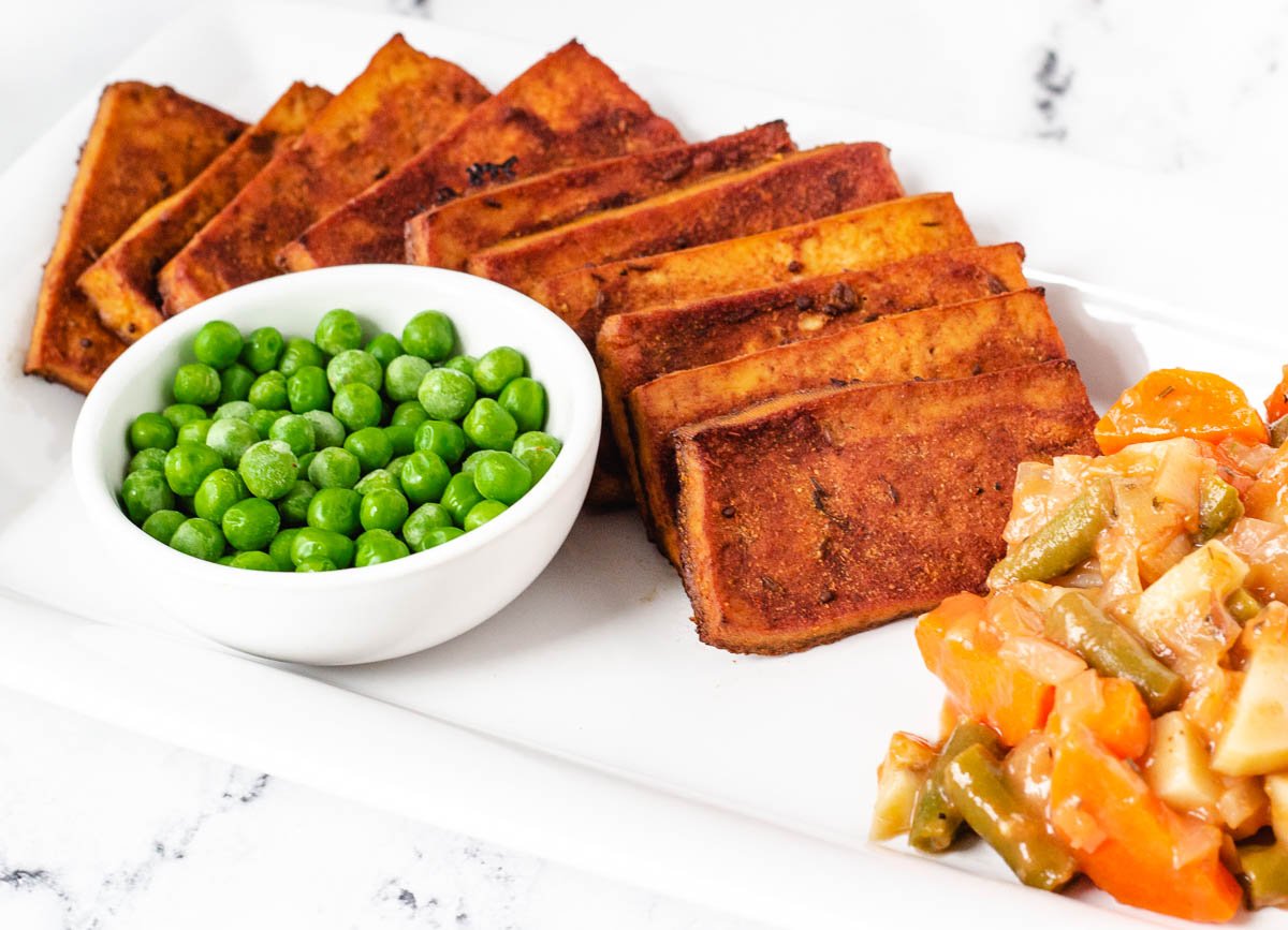 slices of vegan corned beef tofu on white platter with side of peas, potatoes, and carrots