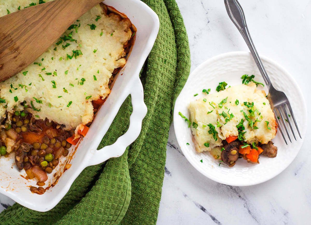 Vegan lentil shepherd's pie in casserole dish with serving taken out, on white plate with fork.