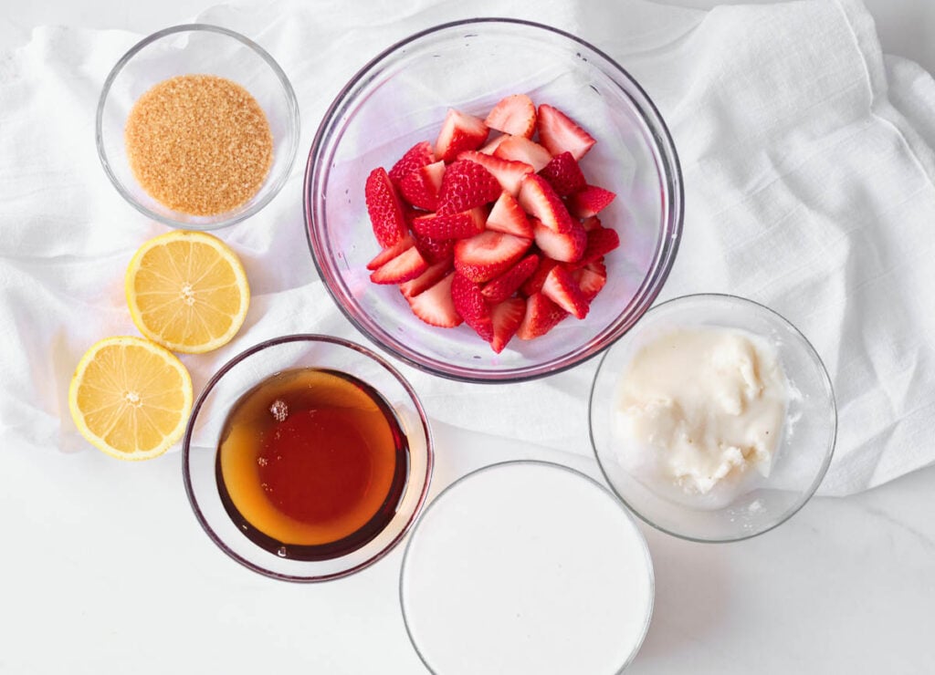 vegan strawberry ice cream ingredients: raw sugar, lemon juice, sliced strawberries, maple syrup, coconut milk, and coconut cream in glass bowls
