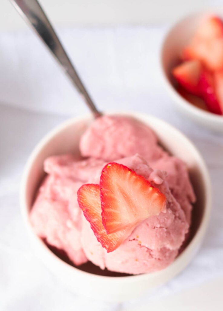 overhead of strawberry ice cream topped with two slices of strawberries in white bowl with spoon
