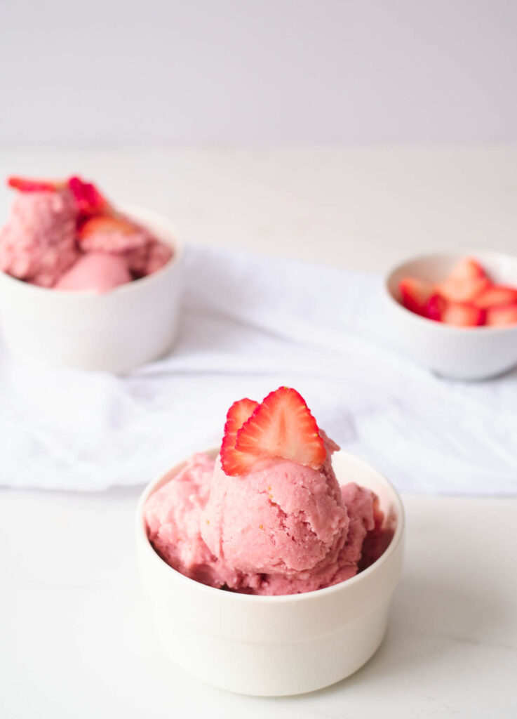 vegan strawberry ice cream in white bowl topped with strawberry slices with slices strawberries and other bowl of ice cream in the background