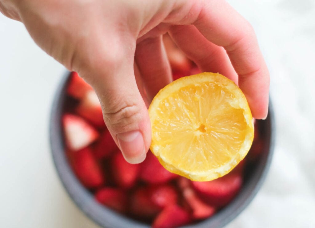 Wedge of lemon, held over pot of fresh strawberries.