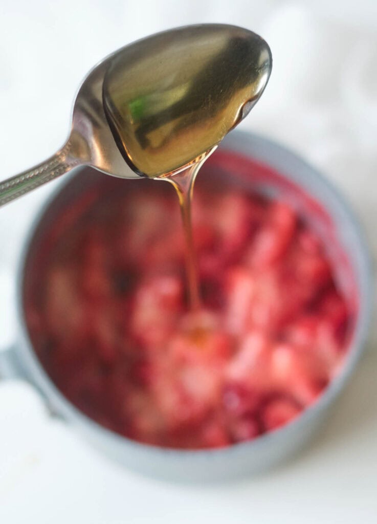 Maple syrup on a spoon poured into pot with simmering strawberries.