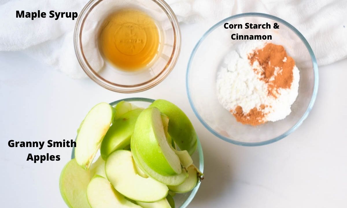 Bowl of sliced granny smith apples, bowl of corn starch and cinnamon, bowl of maple syrup.