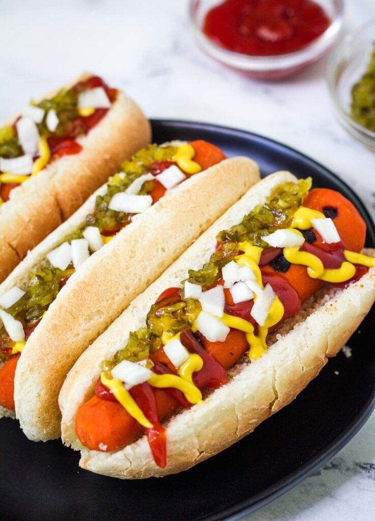 Vegan carrot hot dogs on black plate topped with onion, relish, mustard, and ketchup.
