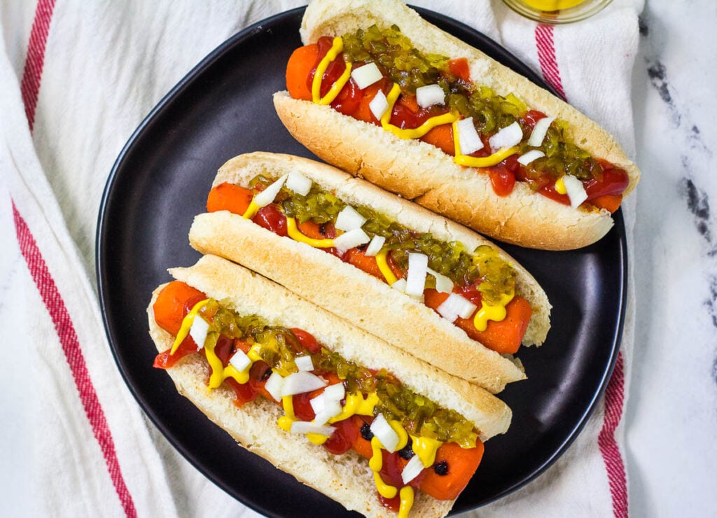 Three carrot hot dogs on black plate topped with relish, onion, mustard, and ketchup.