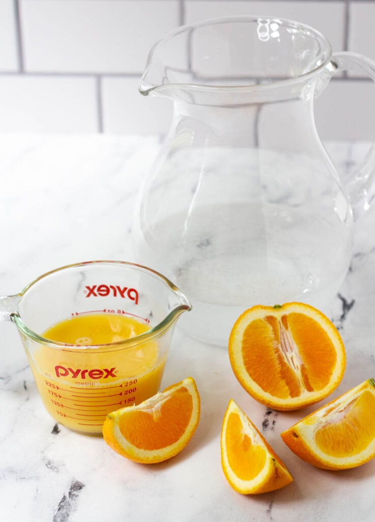 Pitcher of sparkling water, measuring cup filled with orange juice, and an orange cut into slices. 