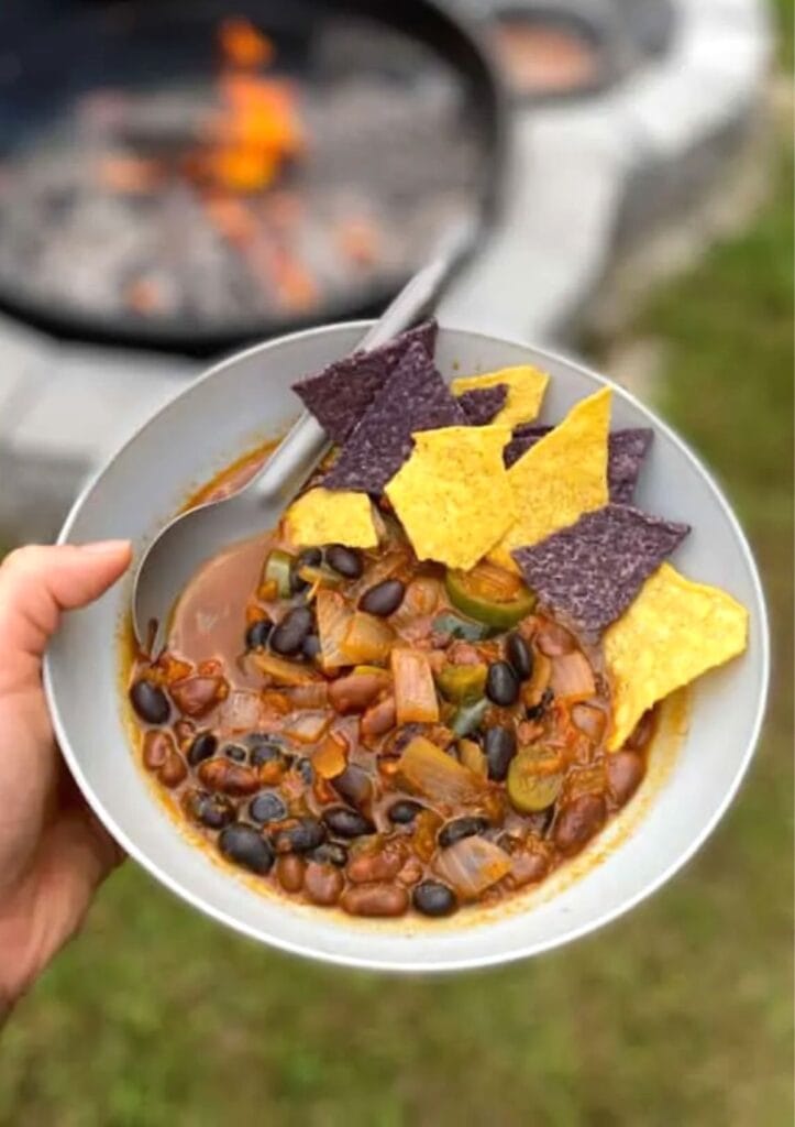 Hand holding a bean chili bowl with chips near a campfire.