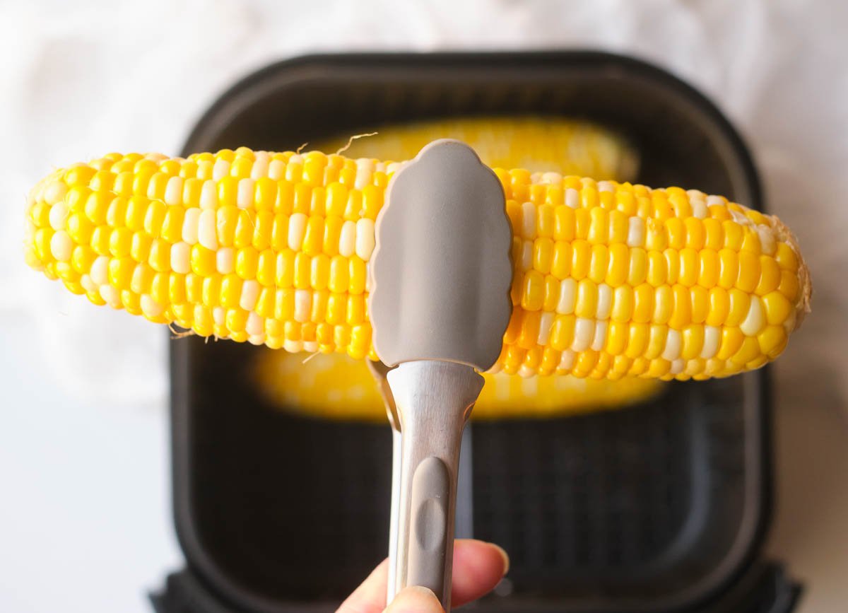 Tongs lifting corn ear out of an air fryer basket.