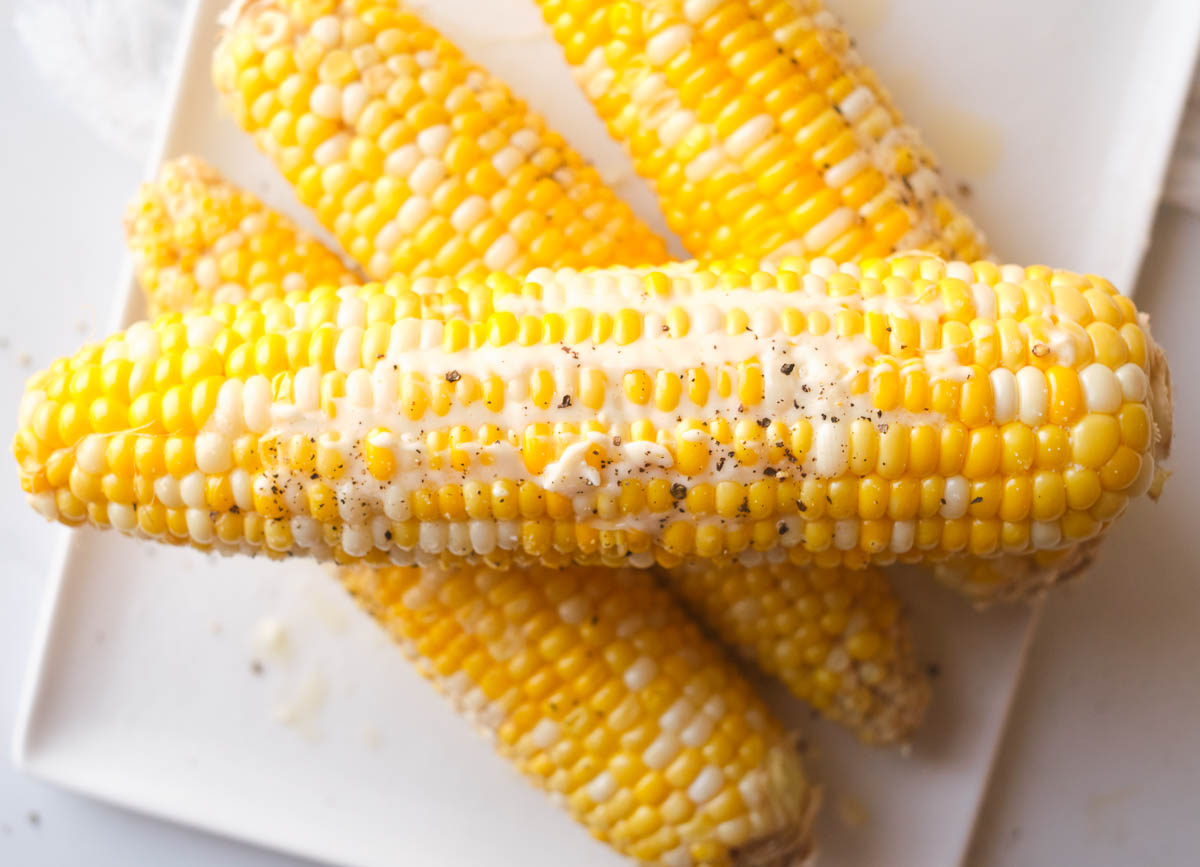 Four cooked ears of corn stacked on a white plate topped with butter, salt, and pepper.