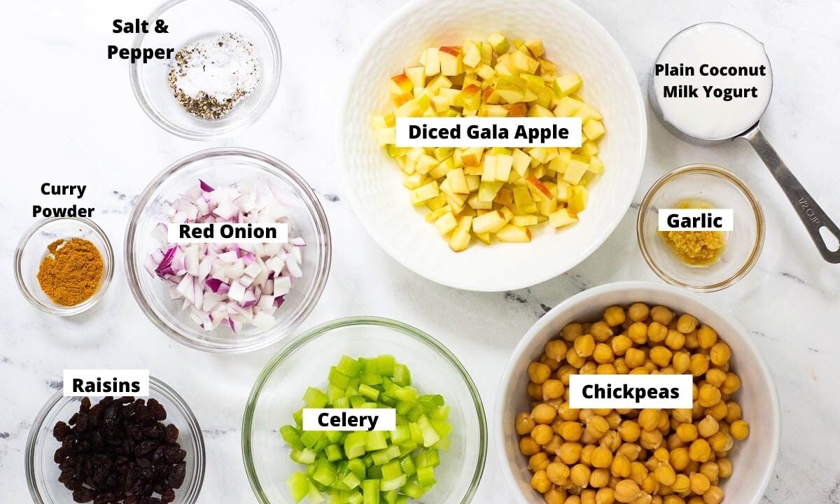 Ingredients for chickpea salad in bowls: salt and pepper, red onion, curry powder, raisins, celery, chickpeas, garlic, diced gala apple, and plain coconut milk yogurt.