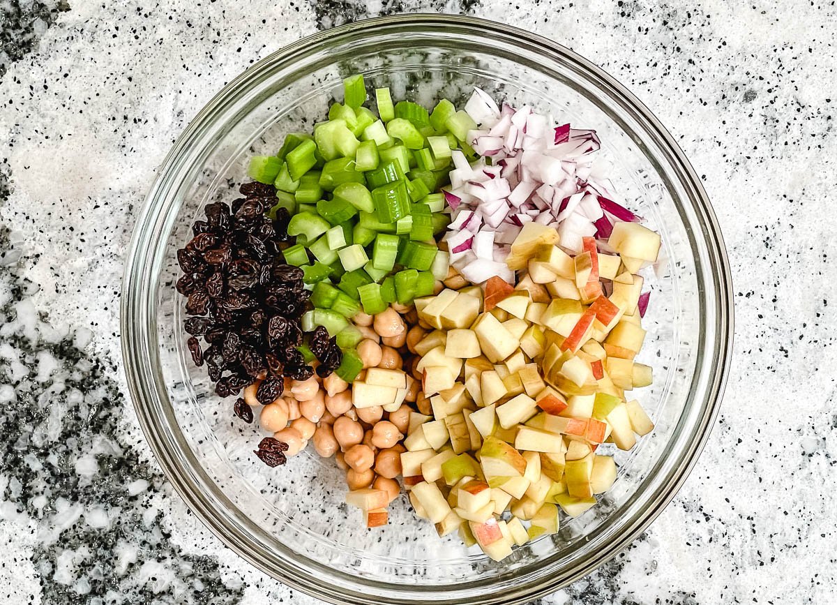 Raisins, diced celery, diced red onion,  diced gala apples, and chickpeas in a glass bowl.