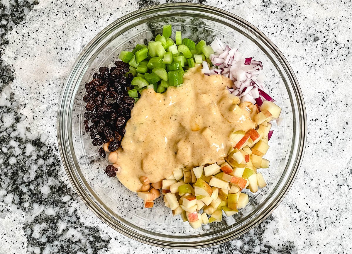 Glass bowl filled with raisins, celery, red onion, apples, and chickpeas covered in a creamy curry dressing.