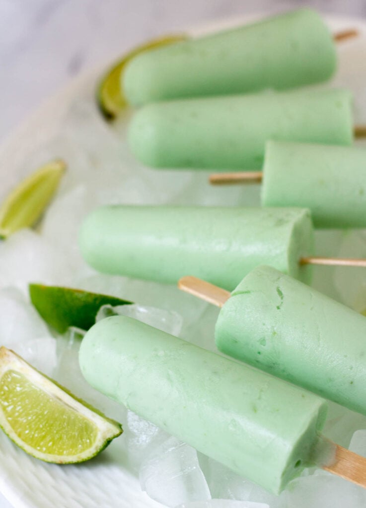 Coconut lime popsicles on white plate with ice and lime wedges.