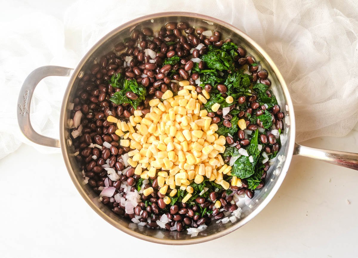 Corn added to black beans, spinach, and onions sautéing in pan.