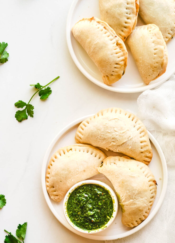Three empanadas on white plate with a side of green chimichurri sauce.