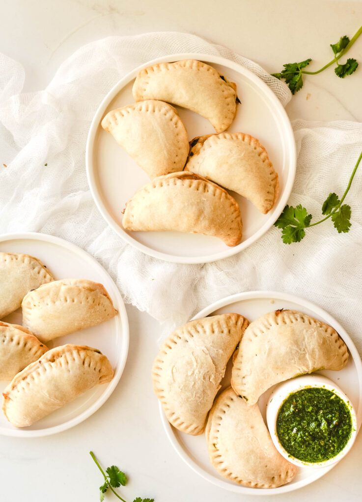 Three empanadas on three white plate with a side of green chimichurri sauce.