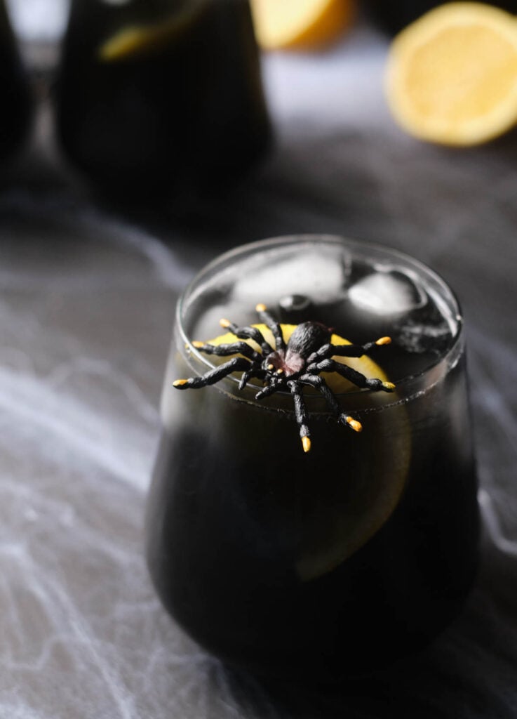 Black widow cocktail in stemless wine glass garnished with lemon and a plastic spider.