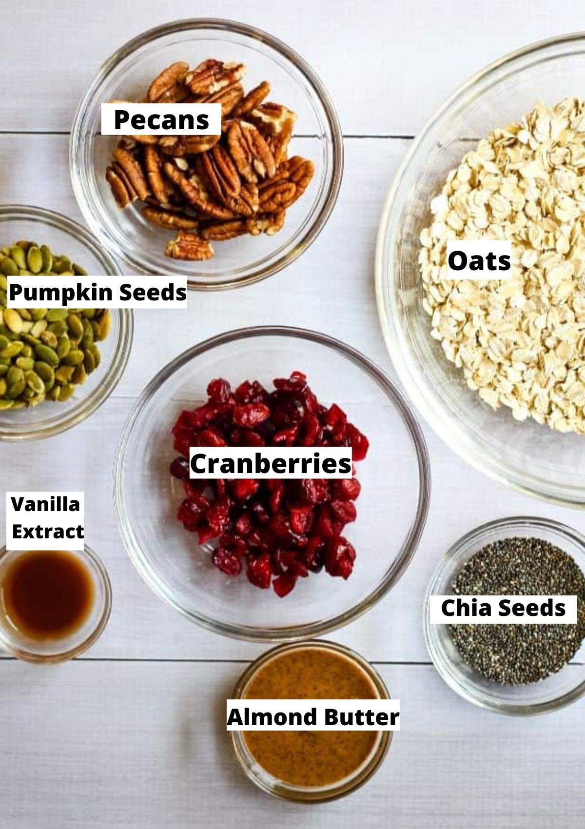 Ingredients for sugar free granola in glass bowls: pecans, oats, pumpkin seeds, cranberries, vanilla extract, almond butter, and chia seeds.
